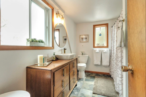A modern bathroom featuring a wooden vanity, a round mirror, a toilet, and a shower curtain, with natural light.