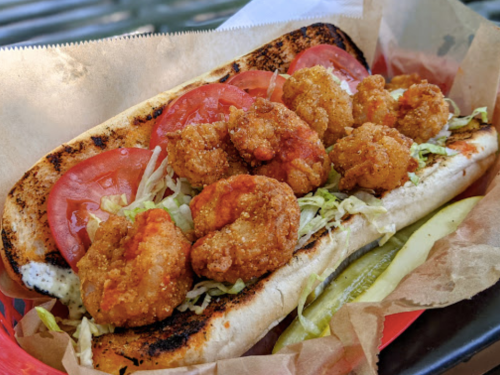 A toasted sandwich filled with crispy shrimp, lettuce, tomatoes, and pickles, served in a red basket.