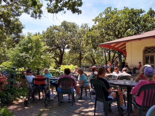 A lively outdoor gathering with people seated at tables under trees, enjoying music and conversation on a sunny day.