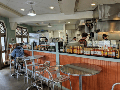 A casual restaurant interior with metal tables, a condiment bar, and staff working in the kitchen area.