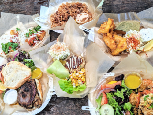 A variety of colorful dishes on paper plates, including seafood, salads, and sandwiches, arranged on a wooden table.
