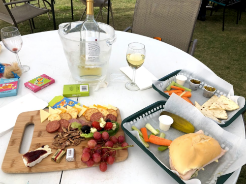 A table with a variety of snacks: cheese, meats, grapes, pickles, carrots, and a sandwich, alongside wine and a bucket.