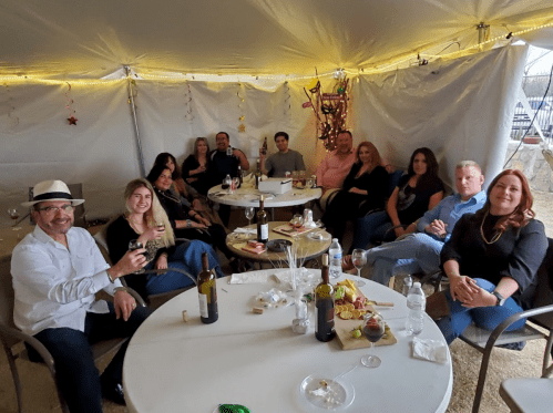 A group of people sitting around tables in a tent, enjoying drinks and snacks, smiling and socializing.