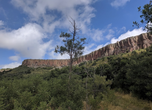 A rocky cliff rises above a lush green landscape under a partly cloudy sky.