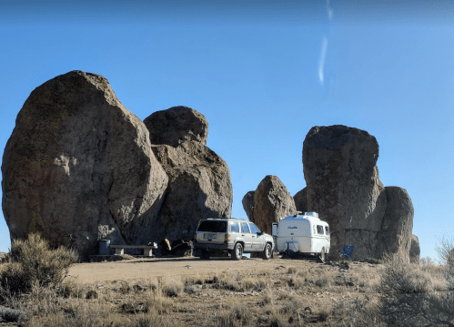 A camper and SUV parked near large boulders in a desert landscape under a clear blue sky.