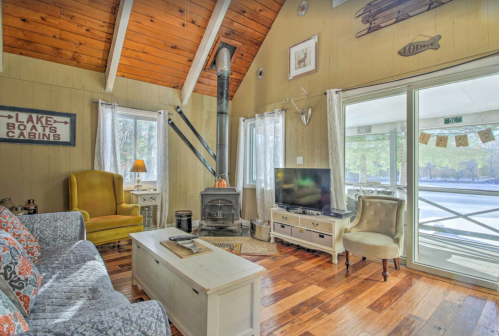 Cozy cabin interior with wooden beams, a stove, comfortable seating, and large windows overlooking nature.