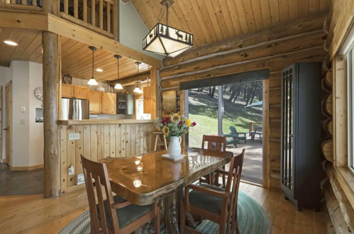 Cozy log cabin interior featuring a wooden dining table, chairs, and a view of the outdoors through large windows.