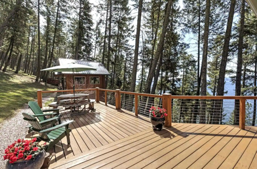 A wooden deck with green chairs and a table, surrounded by trees and overlooking a lake. Bright flowers in pots add color.