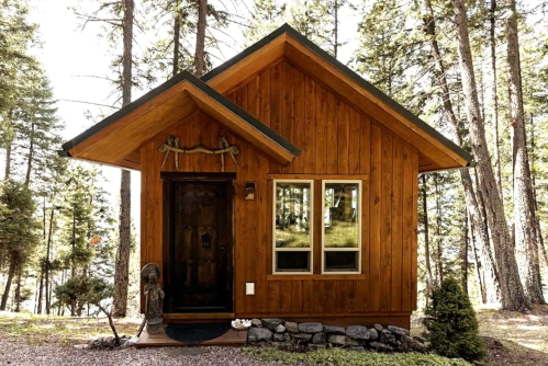 A small wooden cabin surrounded by tall trees, featuring a sloped roof and a welcoming front door.
