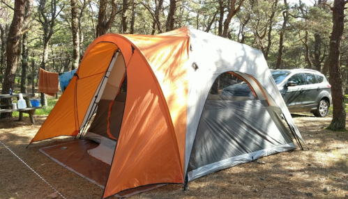 A large orange and gray tent set up in a wooded campsite, with a car parked nearby and trees in the background.
