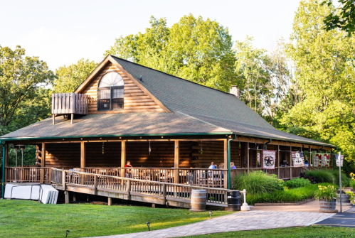 A rustic wooden lodge surrounded by greenery, featuring a spacious porch and large windows.