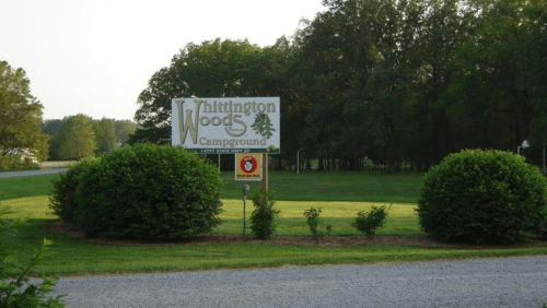 Sign for Whittington Woods Campground surrounded by green bushes and trees, with a gravel path in the foreground.