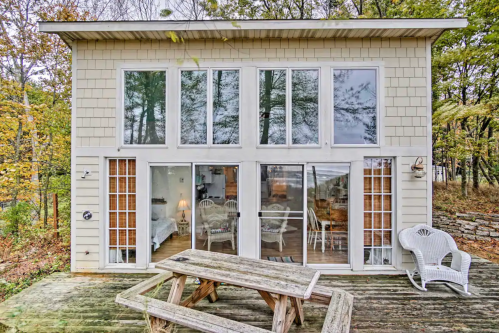 A modern two-story house with large windows, surrounded by trees, featuring a wooden deck and picnic table.