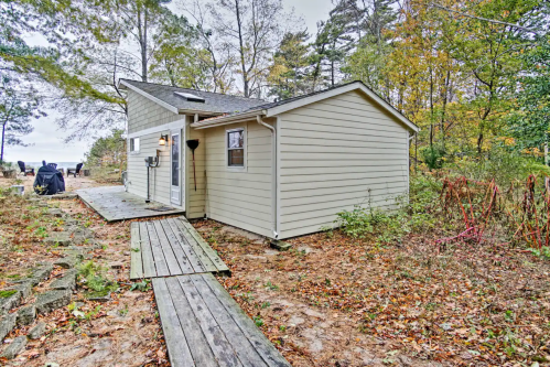 A small, light-colored house surrounded by trees and fallen leaves, with a wooden pathway leading to the entrance.