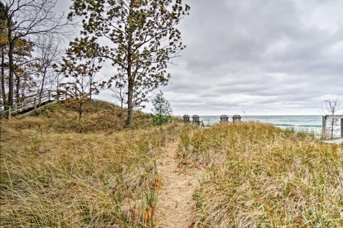 A sandy path through tall grass leads to a cloudy beach with wooden structures near the water's edge.