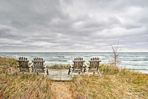Four wooden chairs facing a cloudy ocean, surrounded by tall grass and a wooden deck.