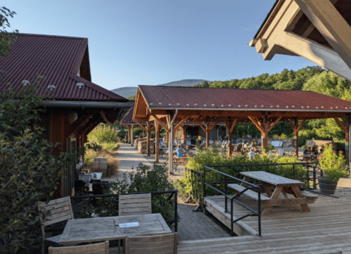 A sunny outdoor dining area with wooden tables and a rustic building, surrounded by greenery and mountains in the background.