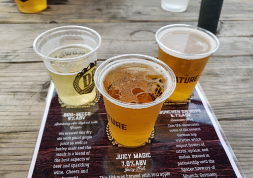 Three cups of beer on a wooden table, with a menu featuring various brews in the background.