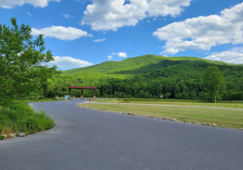 A winding road leads to a green mountain under a blue sky with fluffy clouds, surrounded by lush trees.