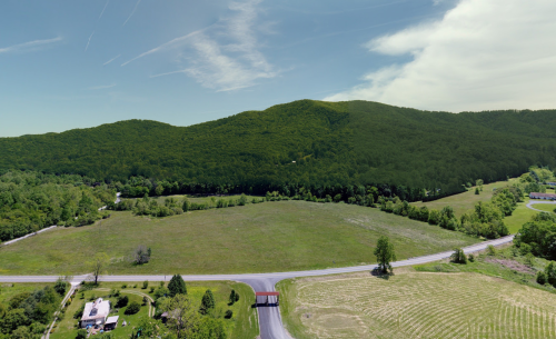 Aerial view of a lush green landscape with rolling hills, fields, and a winding road. Clear blue sky above.