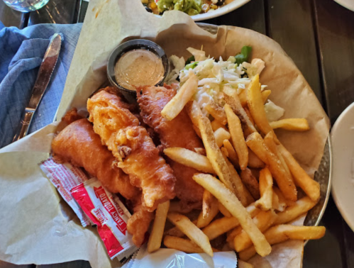 A plate of fish and chips with two battered fish fillets, fries, coleslaw, and dipping sauces.