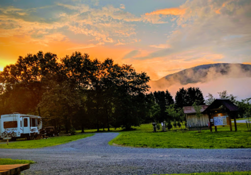 A serene campsite at sunset, with an RV, trees, and misty mountains in the background.