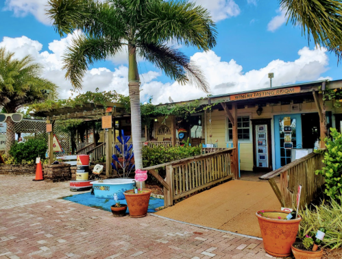 A charming winery tasting room with a wooden deck, potted plants, and palm trees under a blue sky.