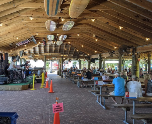 A lively indoor venue with a band performing, surrounded by seated guests at picnic-style tables. Surfboards hang above.