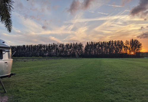 A serene landscape featuring a grassy field, a vintage trailer, and a colorful sunset behind a line of trees.
