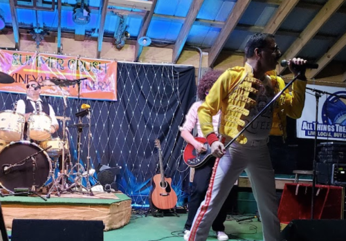 A tribute band performs on stage, featuring a singer in a yellow jacket, a drummer, and a guitarist.