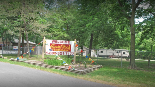 A welcome sign for Crystal Rock Campground, surrounded by greenery and colorful flags, with a trailer visible in the background.