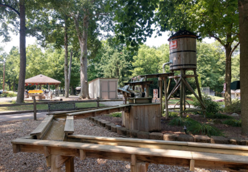 A wooden water play area with a large tank, surrounded by trees and a gazebo in a park setting.