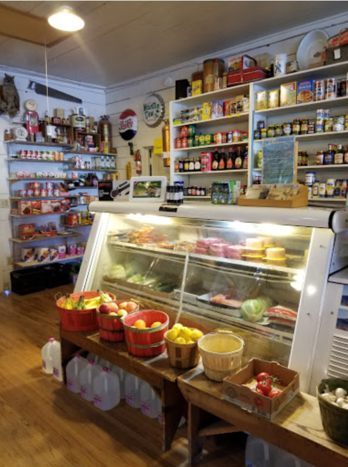 A cozy store interior featuring a display case with meats and cheeses, shelves of canned goods, and baskets of fresh produce.