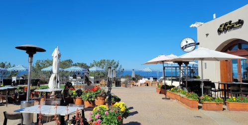 Outdoor dining area with flowers, umbrellas, and a clock, overlooking a clear blue sky and ocean in the background.