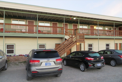 Two-story building with wooden balconies, parked cars in front, and a staircase leading to the upper level.