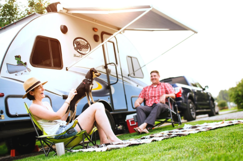 A woman and a man relax outside a camper, enjoying the sun with a small dog, surrounded by lawn chairs and a picnic setup.