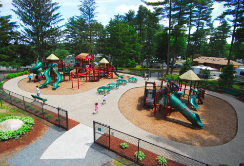 Aerial view of a playground with slides, climbing structures, and children playing, surrounded by trees and a fence.