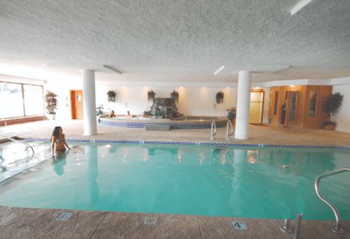 Indoor swimming pool area with a waterfall feature, surrounded by plants and people relaxing.
