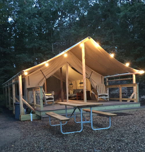 A cozy tent cabin with string lights, wooden deck, and picnic table, surrounded by trees in a serene outdoor setting.