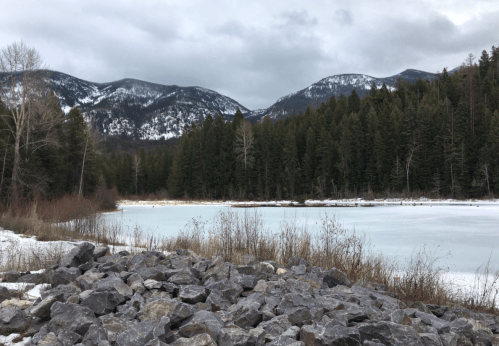 A serene winter landscape featuring a frozen lake, rocky shore, and snow-capped mountains in the background.