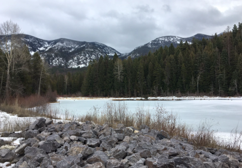 A serene winter landscape featuring a frozen lake, rocky shore, and snow-capped mountains in the background.