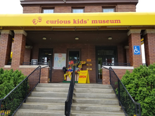 Entrance of the Curious Kids' Museum with yellow awning, stairs, and welcoming signs.