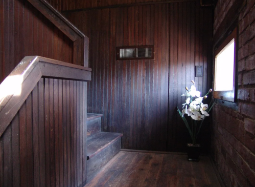 A dimly lit wooden staircase leads to a small window, with a vase of white flowers in the corner.