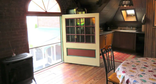 Cozy attic room with wooden floors, a round table, and a kitchen area, featuring a large window and an open door.