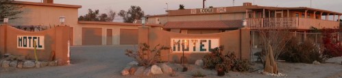 A rustic motel entrance with a sign, surrounded by desert landscaping and a warm sunset glow.