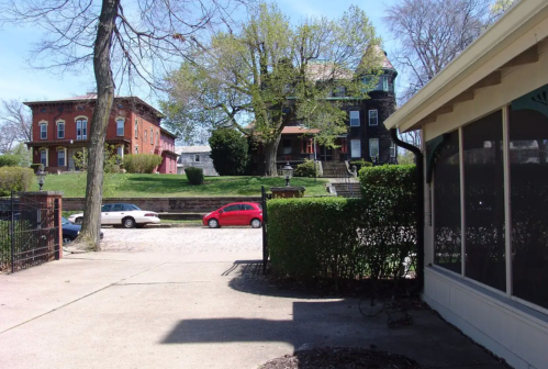 A view of two historic houses across a street, with trees and parked cars in a sunny setting.