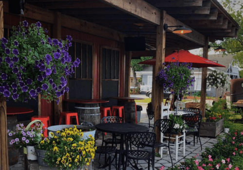 A cozy outdoor seating area with colorful flowers, hanging plants, and tables under a wooden awning.