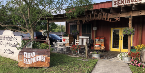 A cozy eatery with a welcoming sign, outdoor seating, and a display of fresh produce and flowers.