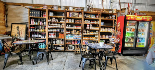 A rustic shop interior with shelves of sauces and snacks, two tables, and a drink cooler in a cozy setting.