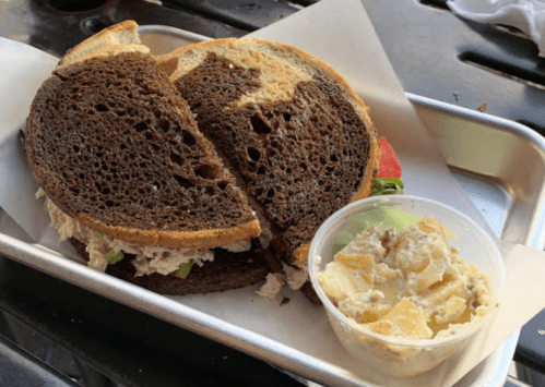 A large sandwich on dark rye bread, filled with chicken salad, served with a side of potato salad in a cup.